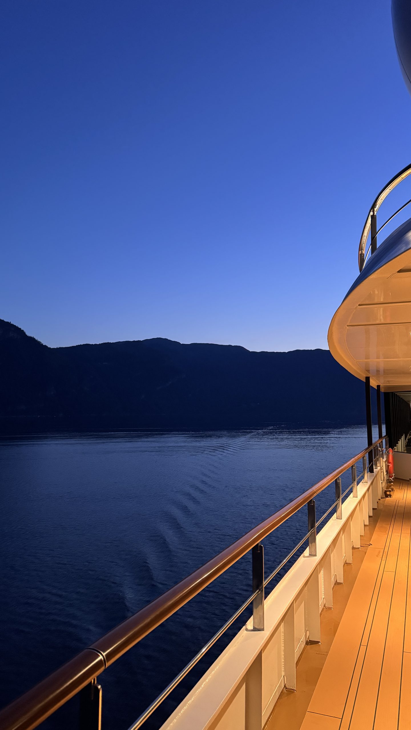 Blue-hour view along the lamplit teak deck of World Navigator, calm fjord and deep blue sky