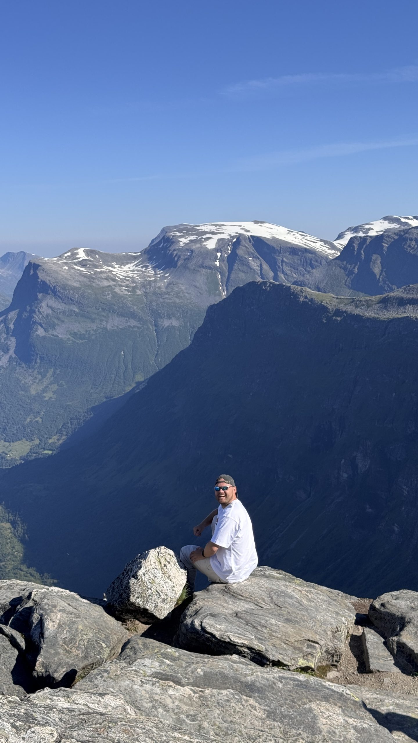 Max Zaharenkov seated on a rocky summit with snow-streaked peaks and a deep valley behind