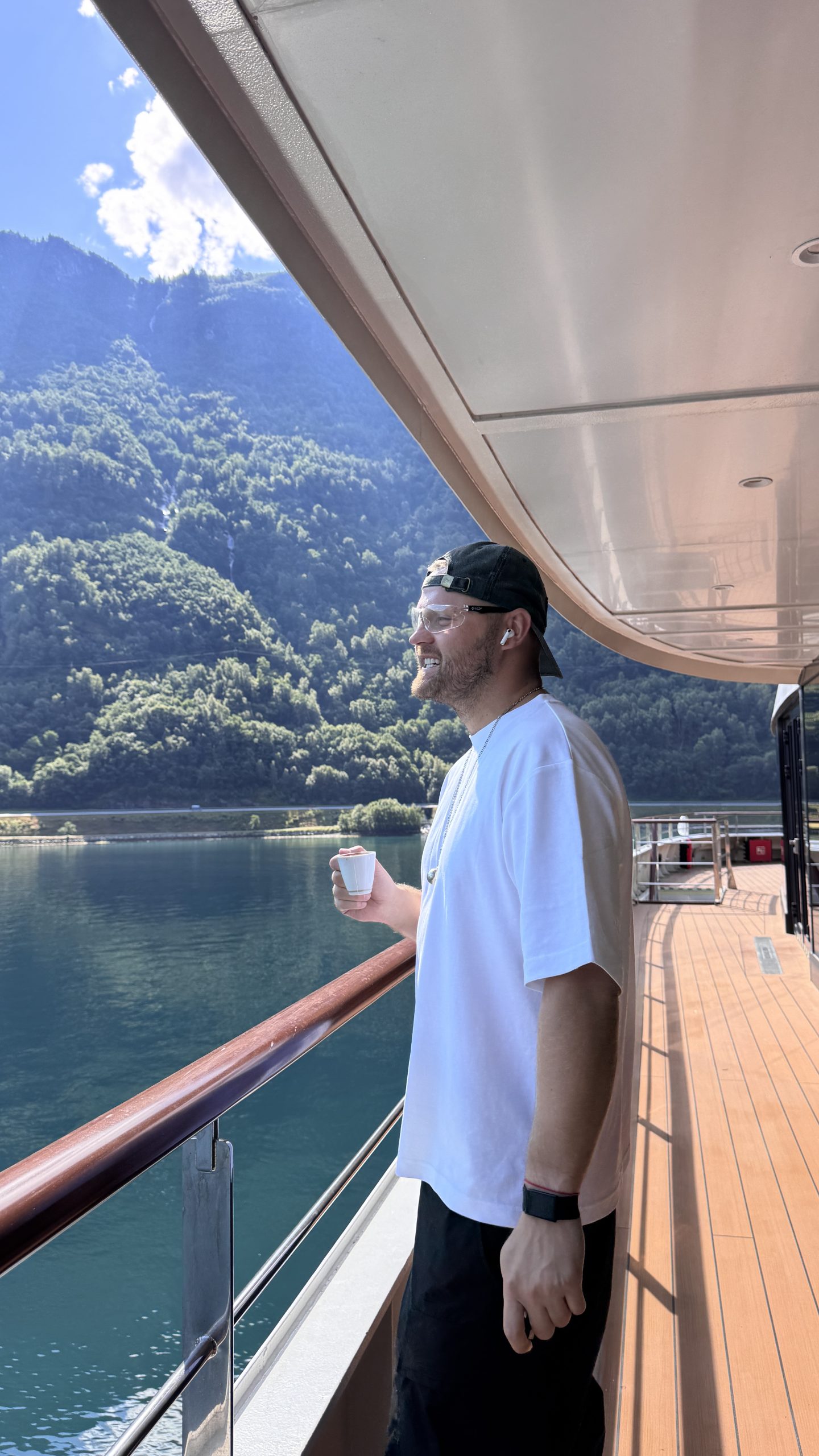 Max Zaharenkov holding a coffee on the teak deck of World Navigator, green fjord wall behind